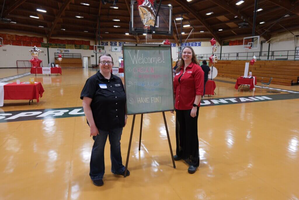 Two smiling women in OTIS shirts standing next to chalkboard in gymnasium.