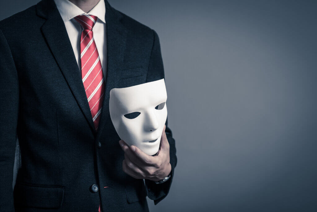Person wearing suit and tie, holding white theater mask.